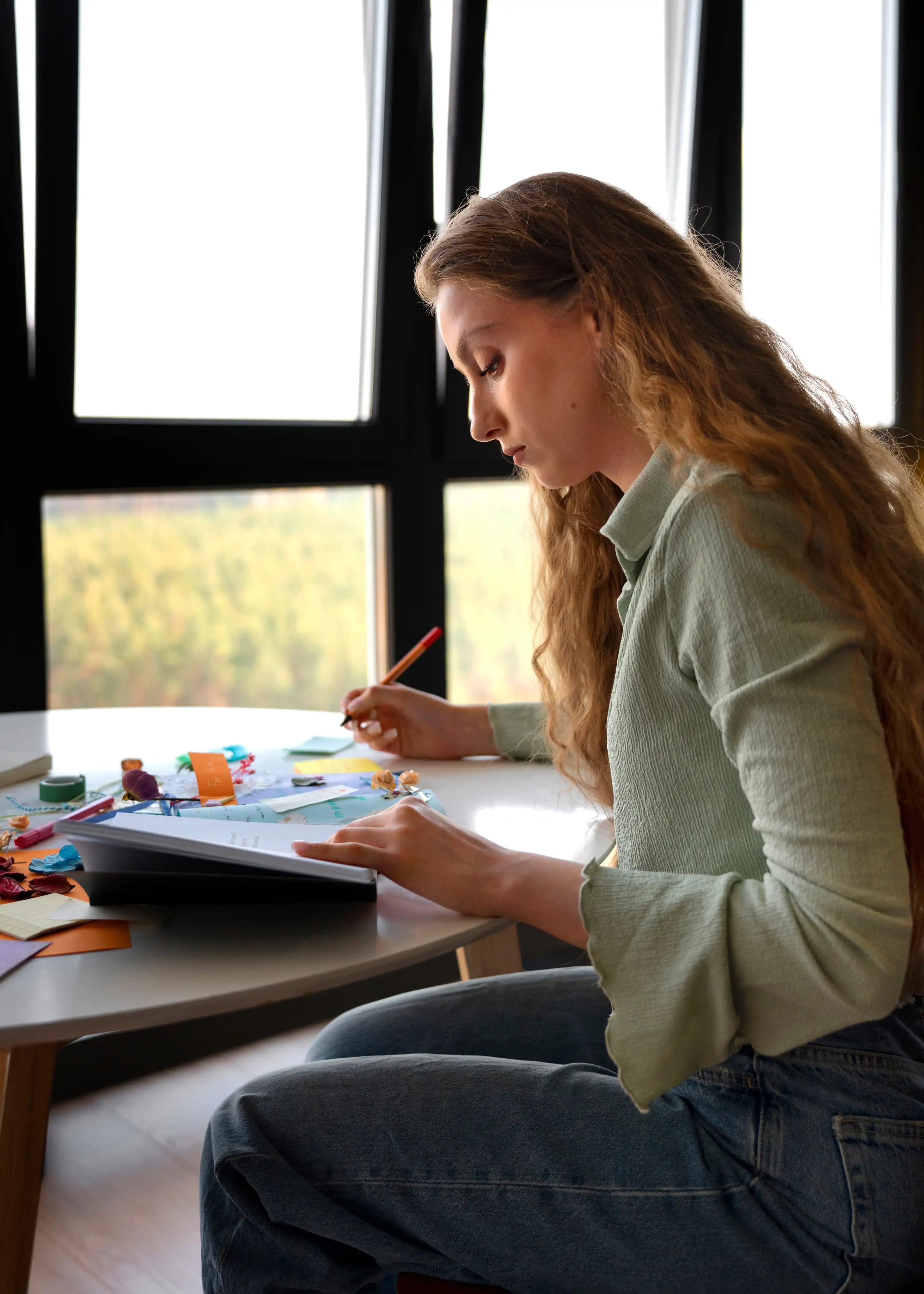 Mulher desenhando a lápis em um caderno perto de uma janela iluminada