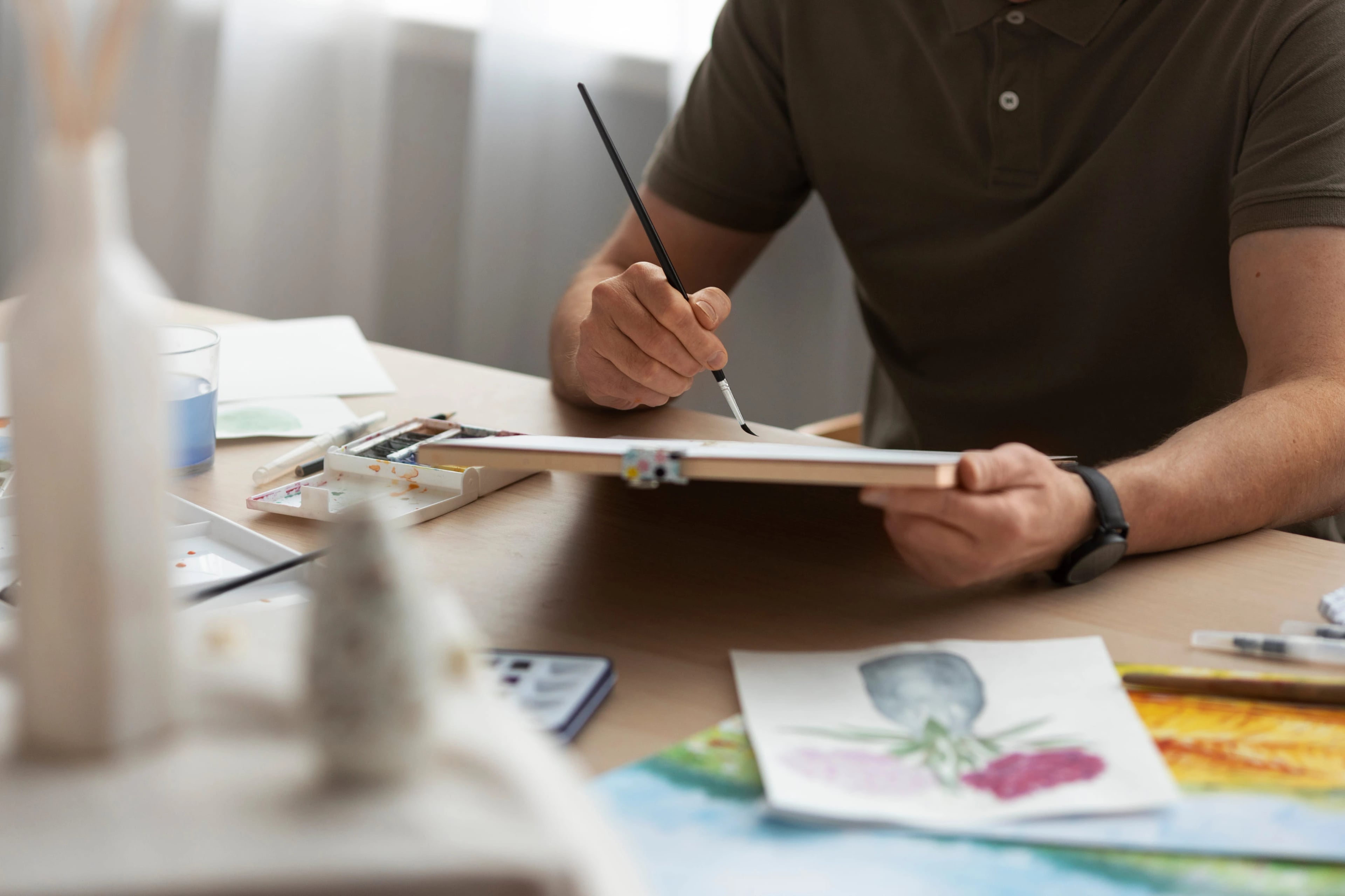 Homem pintando com pincel e aquarela em uma prancheta, à mesa com paleta de tintas, copo de água e estudos de flores