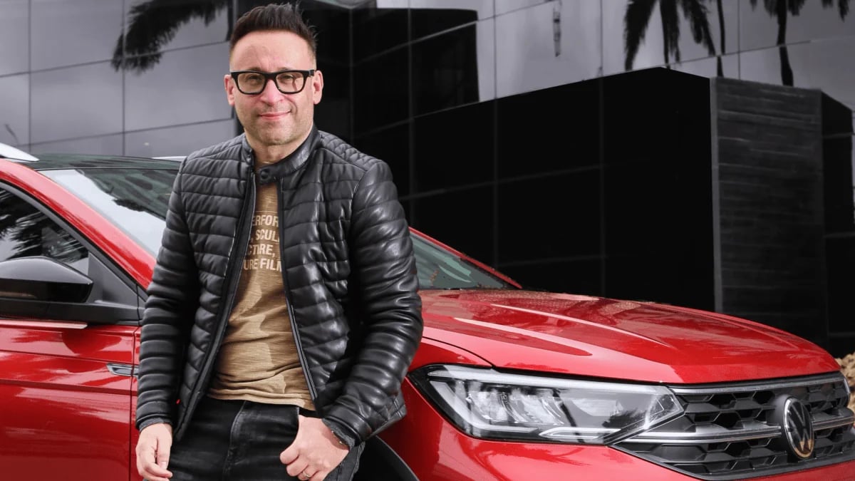 JC Pavone, de jaqueta de couro e óculos, encostado em um carro vermelho