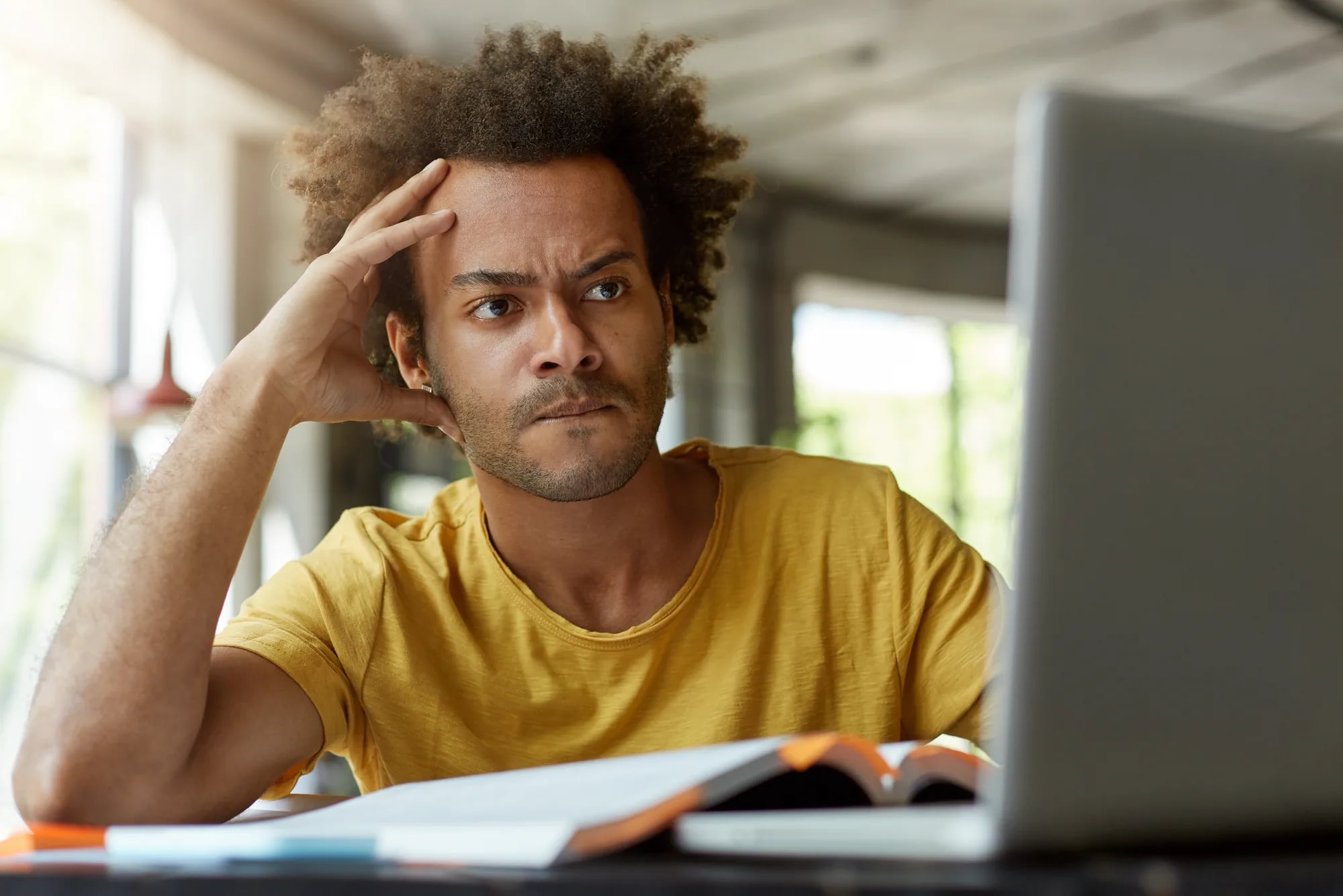 Homem de camiseta amarela com a mão na cabeça, olhando confuso para a tela do notebook