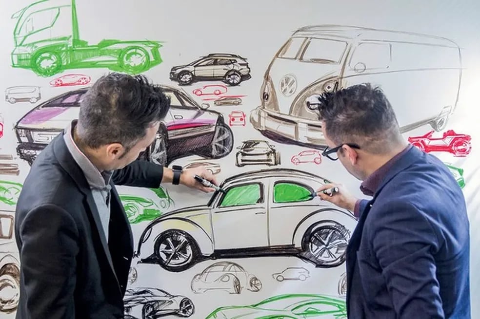 Dois designers desenhando à mão, com marcadores, um Fusca numa parede coberta de sketches de carros