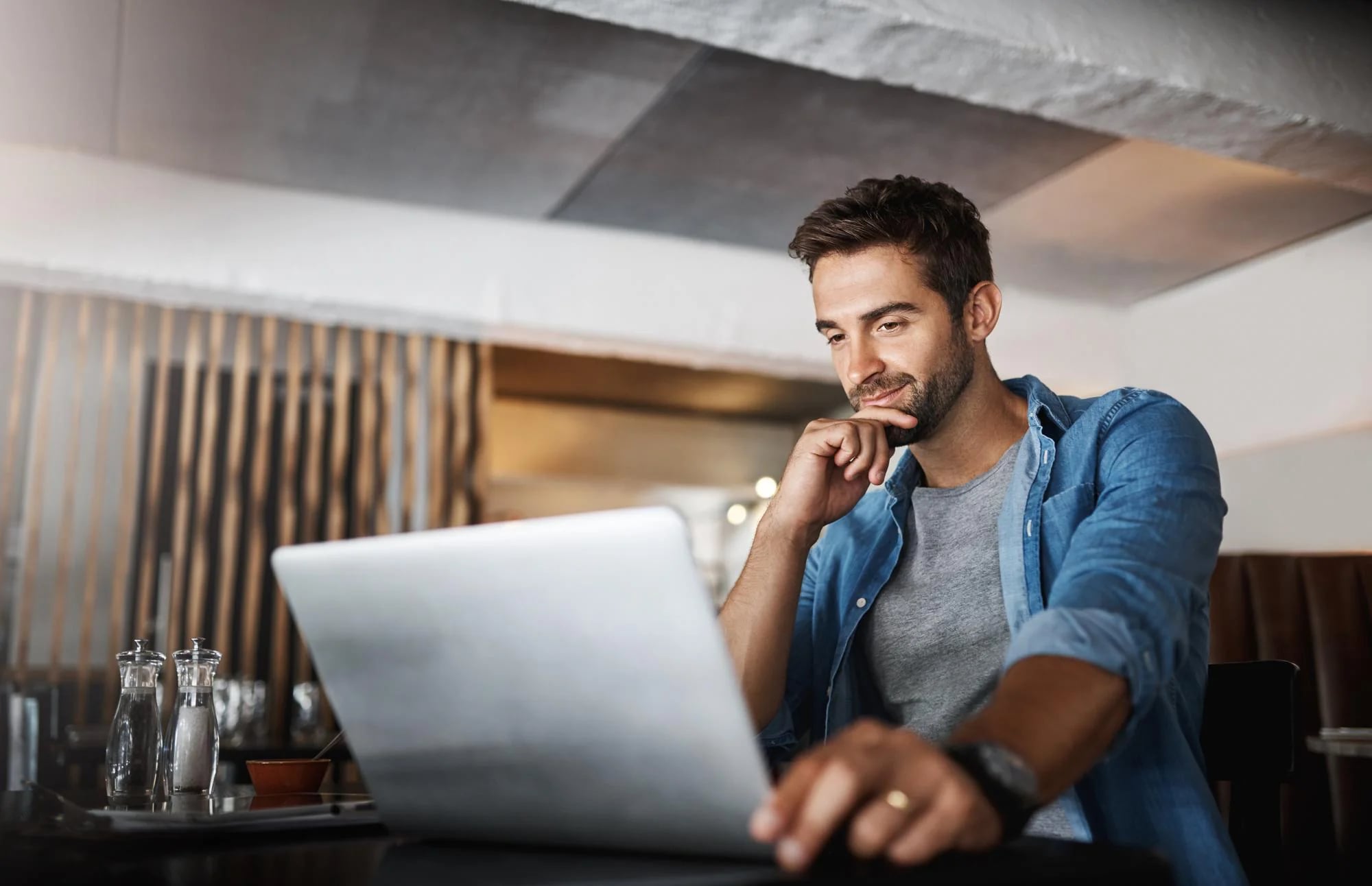 Homem sorrindo com a mão no queixo enquanto usa um notebook numa mesa de café
